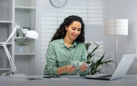 Why-is-Cash-Reconciliation-Important.webp A woman in an office counting cash with a laptop in front of her, representing the importance of financial accuracy and reconciliation.